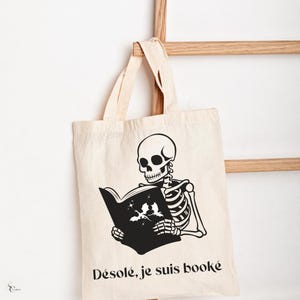 Peut inclure: Un sac tote blanc avec un squelette noir lisant un livre. Le texte sur le sac est "Désolé, je suis booké".