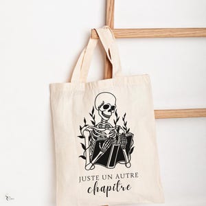 May include: A white tote bag with a black skeleton reading a book. The text on the bag reads "Juste un autre chapitre".