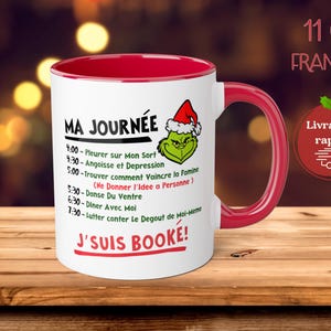 May include: A white ceramic 11 ounce mug with a red rim and handle. The mug features a green Grinch character with a red Santa hat and the text "Ma Journee" and a list of activities. The text "J'suis Booke!" is printed in red at the bottom of the mug.