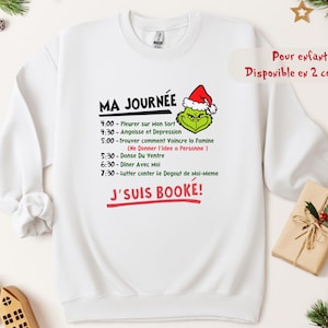 Może przedstawiać: Biała bluza z nadrukiem Grincha w czapce Świętego Mikołaja. Tekst brzmi "MA JOURNÉE" (MÓJ DZIEŃ) z listą czynności. Poniżej listy, tekst "J'SUIS BOOKÉ!" (JESTEM ZAREZERWOWANY!) jest napisany na czerwono. Widoczne są również słowa "Pour enfant! Disponible en 2 couleurs".