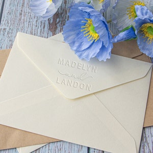 Pu&ograve; includere: Due buste bianche con un'iscrizione in rilievo che recita "Madelyn and Landon" sulla busta superiore. Le buste sono su una superficie di legno blu chiaro con fiori di papavero blu.