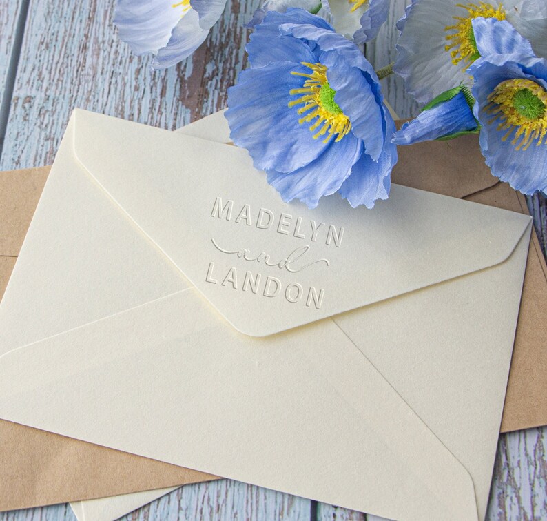 Pu&ograve; includere: Due buste bianche con un'iscrizione in rilievo che recita "Madelyn and Landon" sulla busta superiore. Le buste sono su una superficie di legno blu chiaro con fiori di papavero blu.