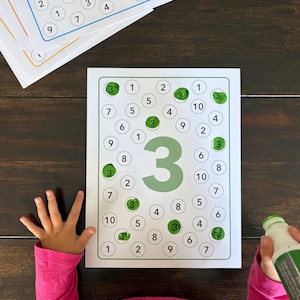 May include: Educational activity sheets with the number 3 highlighted, featuring circles with numbers 1-10. A green marker and a green magnetic ball are visible. A child's hand is also visible.