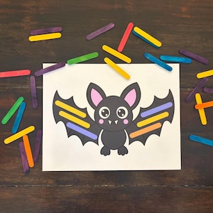 May include: A white rectangular card featuring a cartoon bat illustration with large eyes, pink ears, and cheeks. The bat's wings are decorated with colorful craft sticks in yellow, orange, purple, and blue. Additional loose craft sticks surround the card.