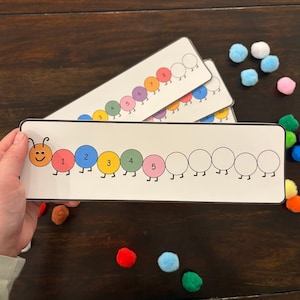 May include: Educational counting cards featuring a colorful caterpillar design with numbered circles. The cards are white with a black border and are accompanied by colorful pom-poms. The caterpillar has a smiling face and is numbered 1 through 5.