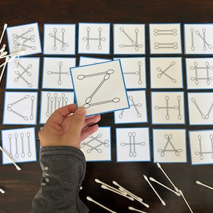 Könnte beinhalten: Eine Hand hält eine Karte mit einem 'Z' aus Wattestäbchen, umgeben von anderen Karten mit verschiedenen Wattestäbchenanordnungen. Die Karten haben einen blauen Rand. Wattestäbchen sind auf einer dunklen Holzoberfläche verstreut.