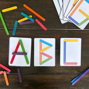 May include: Wooden craft sticks in various colors are arranged to form the letters A, B, and C on white cards. Additional sticks and cards are scattered on a dark wooden surface, creating a colorful educational display.