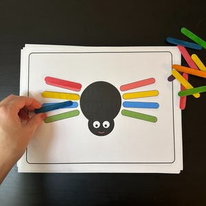 Könnte beinhalten: Ein schwarzes und weißes Spinnenbastelwerk mit einem lächelnden Gesicht. Die Spinne hat acht Beine aus bunten Eis am Stiel-Stäbchen. Die Stäbchen sind rot, gelb, blau und grün.