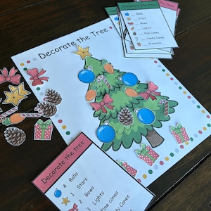 May include: A Christmas-themed activity set with a decorated tree illustration and cut-out decorations. The set includes balls, stars, bows, lights, pine cones, candy canes, and presents. The words "Decorate the Tree" are printed on the page.
