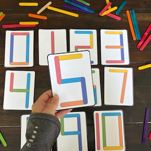 May include: Educational flashcards with numbers 0-9, each number constructed from colorful popsicle sticks. A hand holds the number 5 card. Additional sticks in various colors are scattered around the cards on a dark wooden surface.