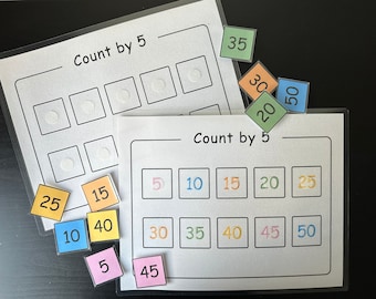 Puede incluir: Dos hojas blancas laminadas con el texto "Contar de 5 en 5" y los números 5, 10, 15, 20, 25, 30, 35, 40, 45 y 50 impresos en diferentes colores. Hay cuadrados coloridos con números en ellos, algunos de los cuales están colocados en las hojas.