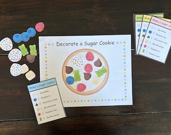 May include: A children's activity set featuring a paper cookie with various toppings, including sprinkles, blueberries, chocolate chips, marshmallows, raspberries, and gummy bears. The set includes cards with instructions. The text "Decorate a Sugar Cookie" is visible.