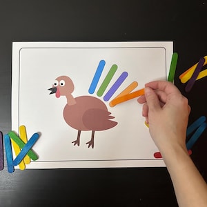 May include: A brown turkey with a rainbow tail made of colorful craft sticks. The turkey is on a white sheet of paper. There are extra craft sticks in different colors on the table.
