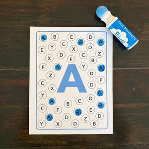 Op de afbeelding: Een wit werkblad met een grote lichtblauwe letter A in het midden, omringd door verschillende letters in cirkels. Verschillende cirkels met de letter A zijn gevuld met blauwe inkt. Een blauwe dot marker ligt ernaast.