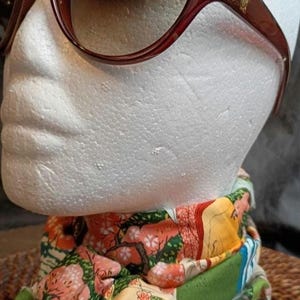 May include: Burgundy-colored sunglasses with a gold emblem on the temple. A colorful scarf with a floral and landscape design is draped below the sunglasses. The scarf features shades of pink, green, and yellow.