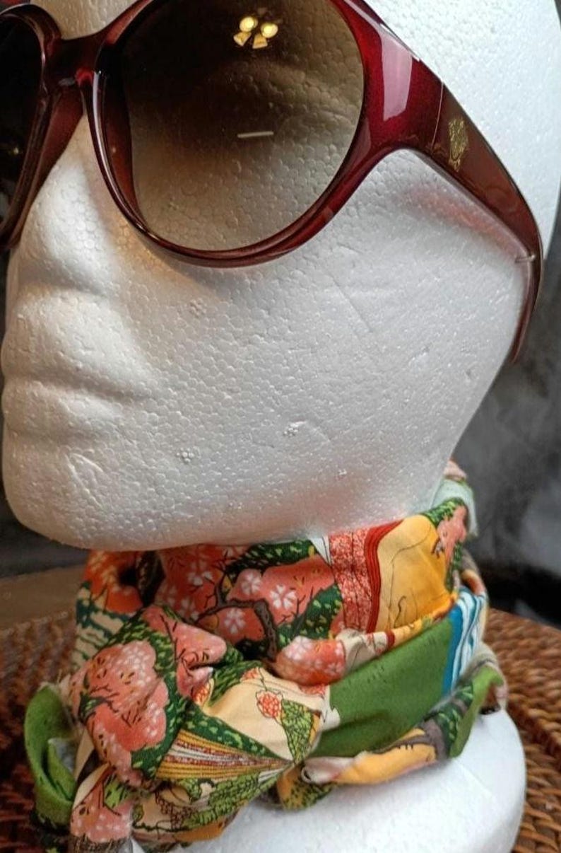 May include: Burgundy-colored sunglasses with a gold emblem on the temple. A colorful scarf with a floral and landscape design is draped below the sunglasses. The scarf features shades of pink, green, and yellow.