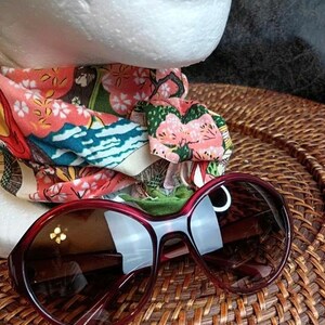 May include: Burgundy-framed sunglasses with dark lenses rest on a woven surface. A colorful patterned headband with floral and landscape motifs is partially visible. The sunglasses have a rounded shape and a thick frame.
