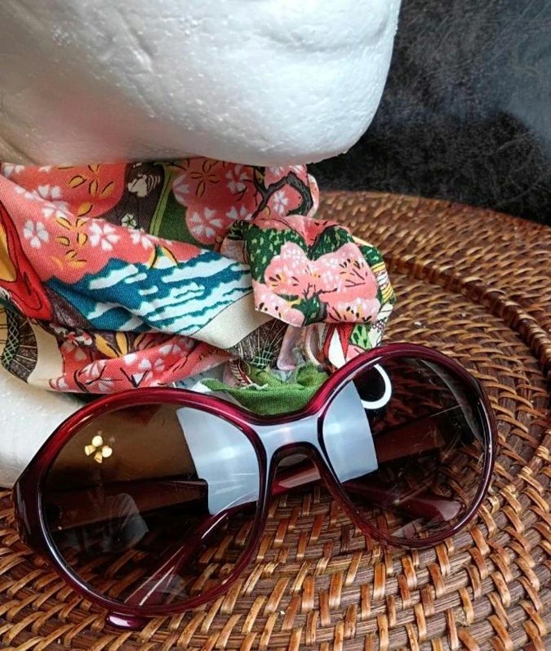 May include: Burgundy-framed sunglasses with dark lenses rest on a woven surface. A colorful patterned headband with floral and landscape motifs is partially visible. The sunglasses have a rounded shape and a thick frame.