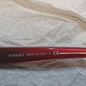 May include: Close-up of a red Versace eyeglass frame. The text "VERSACE MADE IN ITALY - C CE" is printed in white on the frame. The frame is set against a textured, off-white background.