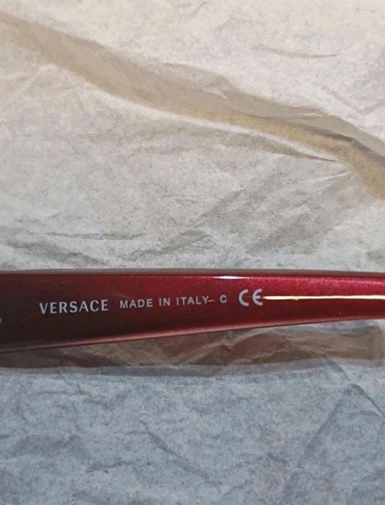 May include: Close-up of a red Versace eyeglass frame. The text "VERSACE MADE IN ITALY - C CE" is printed in white on the frame. The frame is set against a textured, off-white background.