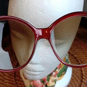 May include: Large, round, burgundy-colored sunglasses with tinted lenses. The frames are thick and have a glossy finish. The sunglasses are set against a white background.