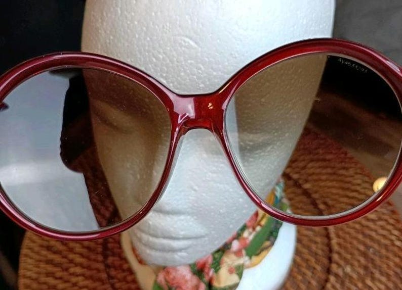 May include: Large, round, burgundy-colored sunglasses with tinted lenses. The frames are thick and have a glossy finish. The sunglasses are set against a white background.