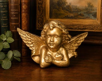 May include: A gold-colored cherub figurine featuring a winged angel with curly hair and arms crossed over its chest. The sculpture has a metallic finish and displays detailed feather patterns on the wings, capturing a classic bust-style design.