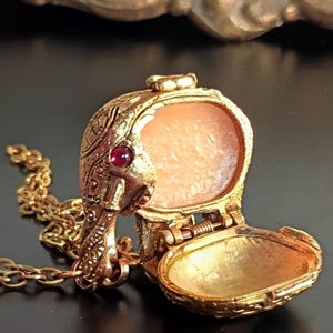 May include: A gold-colored locket necklace with an ornate design. The locket is open, revealing a light-colored interior. A small, red gemstone is set into the side of the locket. The necklace chain is also gold-colored.
