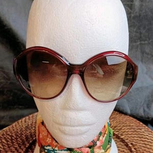 May include: Large, burgundy-framed sunglasses with gradient lenses are displayed on a white mannequin head. The sunglasses have a rounded, oversized shape. A colorful floral scarf is draped around the mannequin's neck.