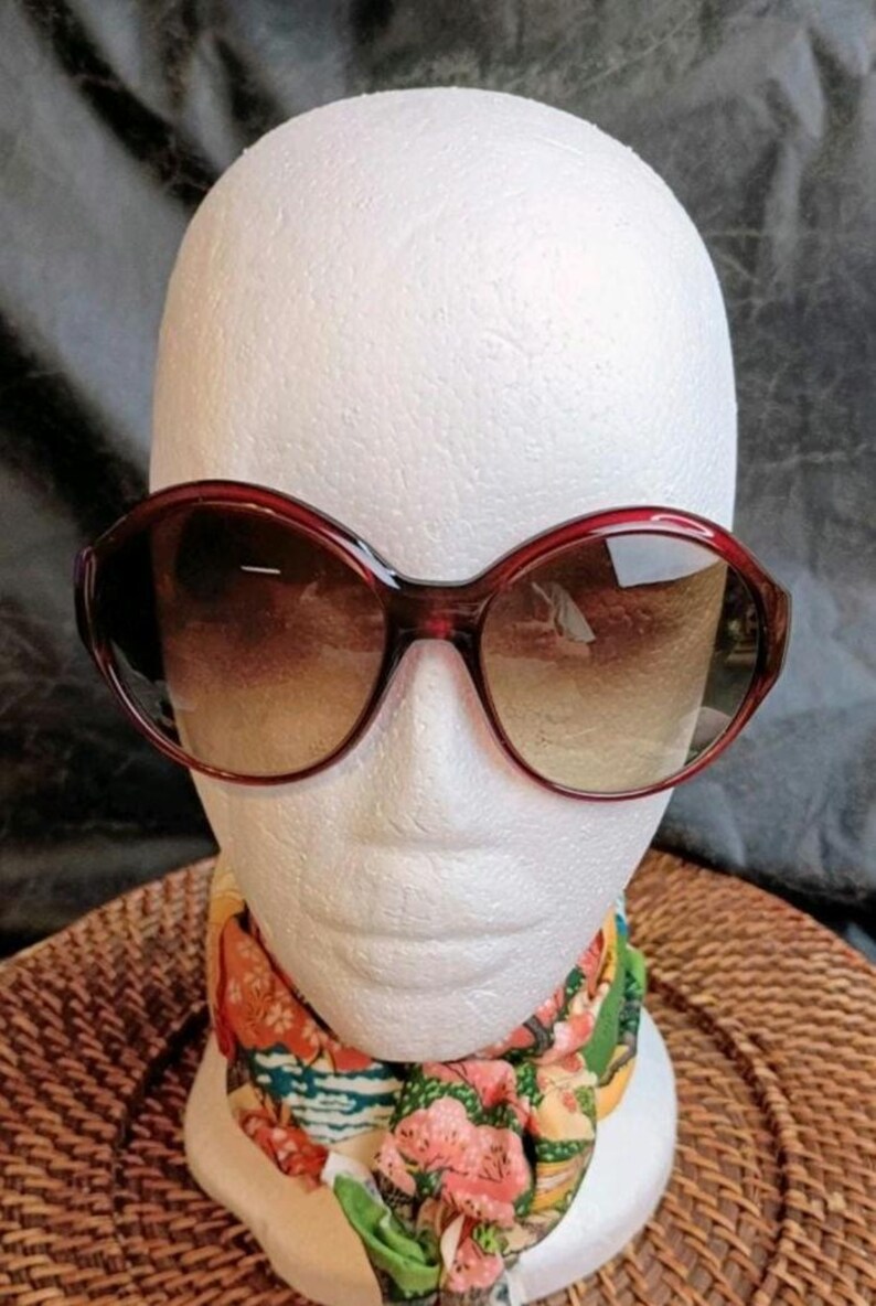 May include: Large, burgundy-framed sunglasses with gradient lenses are displayed on a white mannequin head. The sunglasses have a rounded, oversized shape. A colorful floral scarf is draped around the mannequin's neck.