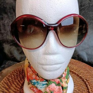May include: Large, dark red-framed sunglasses with gradient lenses. The sunglasses are displayed on a white mannequin head, with a colorful scarf around the neck. The frames have a rounded, oversized shape.