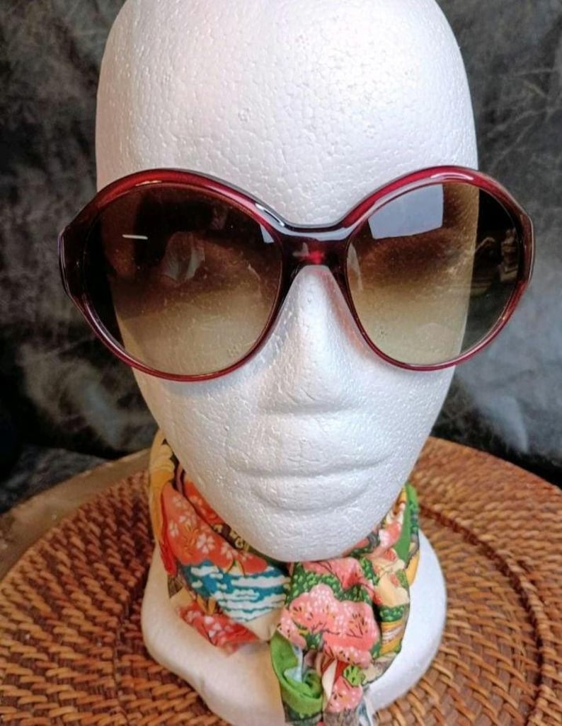 May include: Large, dark red-framed sunglasses with gradient lenses. The sunglasses are displayed on a white mannequin head, with a colorful scarf around the neck. The frames have a rounded, oversized shape.