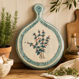 May include: A round ceramic serving board with a handle, featuring a cream-colored center with a hand-painted blue and pink floral sprig. The border is light blue with black cursive text listing herb names including Basilico, Salvia, and Rosmarino.