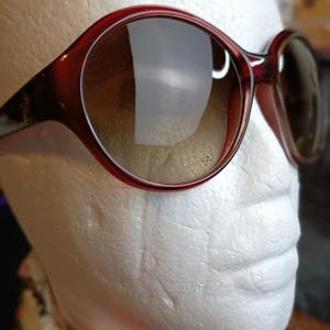 May include: Red-brown sunglasses with large, rounded frames and gradient lenses. The sunglasses are set against a white background, showcasing their shape and color. The lenses transition from a darker shade at the top to a lighter shade at the bottom.