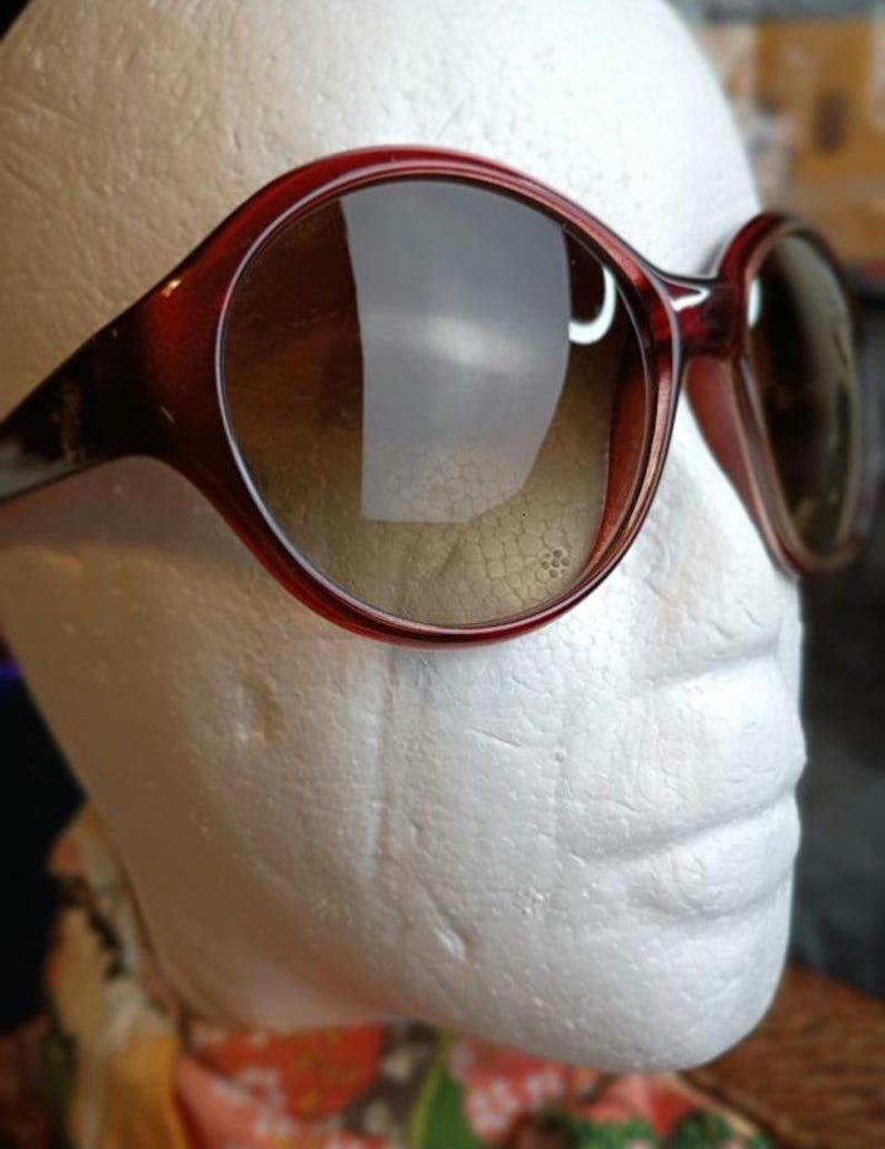 May include: Red-brown sunglasses with large, rounded frames and gradient lenses. The sunglasses are set against a white background, showcasing their shape and color. The lenses transition from a darker shade at the top to a lighter shade at the bottom.