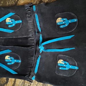 May include: Black leather chaps with turquoise accents. Features include turquoise cactus appliques, fringe, and turquoise ties. The chaps have silver-colored metal accents.