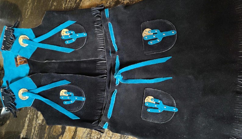 May include: Black leather chaps with turquoise accents. Features include turquoise cactus appliques, fringe, and turquoise ties. The chaps have silver-colored metal accents.