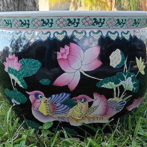 May include: A large, round black ceramic planter with a detailed floral and bird design. The planter features pink lotus flowers, green leaves, and colorful birds. The rim has a decorative border with pink and green accents.
