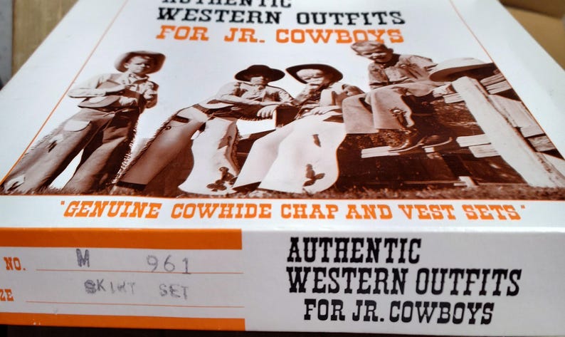 May include: A box featuring a sepia-toned image of children in Western outfits. The box reads "AUTHENTIC WESTERN OUTFITS FOR JR. COWBOYS" and "GENUINE COWHIDE CHAP AND VEST SETS". The box also has a size and style label.