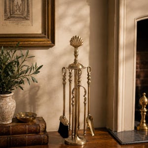 May include: A brass fireplace tool set featuring a central stand topped with a shell-shaped finial. The set includes a poker, a shovel, and a brush with black bristles, all suspended from a circular frame with a weighted base and polished finish.