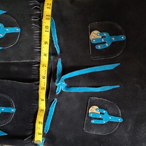 May include: Black suede chaps with turquoise accents and cactus appliqu&eacute;s. The chaps measure approximately 12 inches in length. Features include fringe detailing and decorative stitching, showcasing a Western-inspired design.