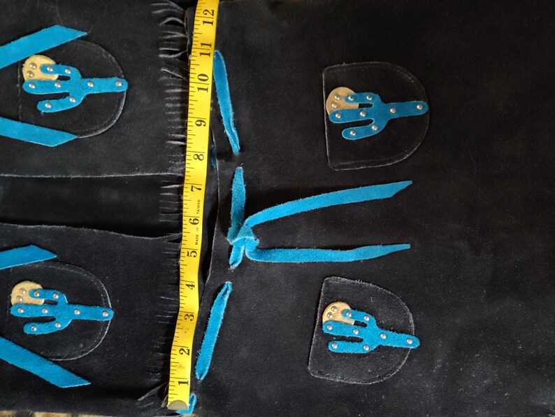 May include: Black suede chaps with turquoise accents and cactus appliqu&eacute;s. The chaps measure approximately 12 inches in length. Features include fringe detailing and decorative stitching, showcasing a Western-inspired design.