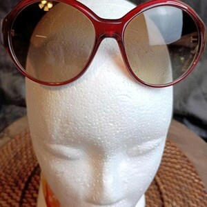 May include: Red-framed sunglasses with oval lenses. The sunglasses are displayed on a white mannequin head. The lenses have a gradient tint, transitioning from a darker shade at the top to a lighter shade at the bottom.