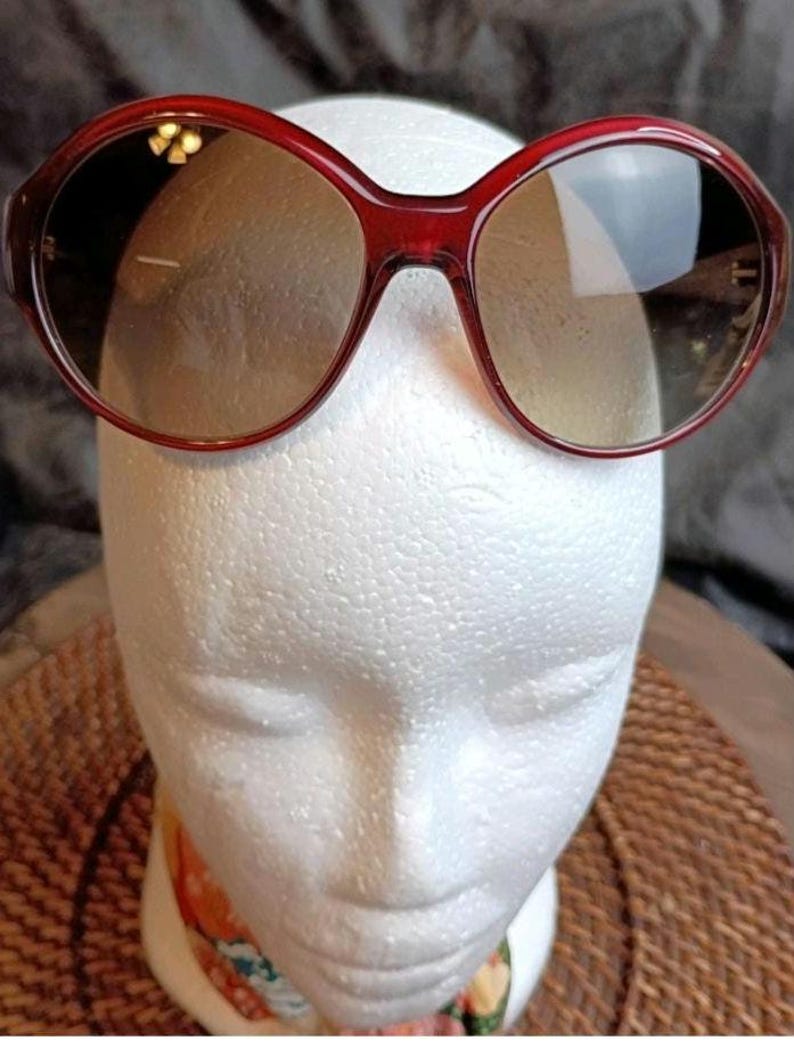 May include: Red-framed sunglasses with oval lenses. The sunglasses are displayed on a white mannequin head. The lenses have a gradient tint, transitioning from a darker shade at the top to a lighter shade at the bottom.