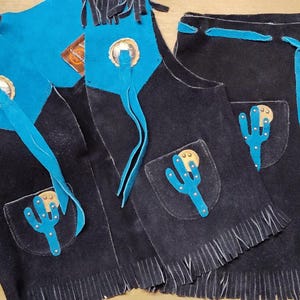 May include: A set of black suede cowboy chaps and vest with turquoise accents and fringe. The chaps and vest feature cactus and medallion details. A book titled "Authentic Western Outfits for Jr. Cowboys" is also in the image.