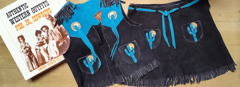May include: A set of black suede cowboy chaps and vest with turquoise accents and fringe. The chaps and vest feature cactus and medallion details. A book titled "Authentic Western Outfits for Jr. Cowboys" is also in the image.
