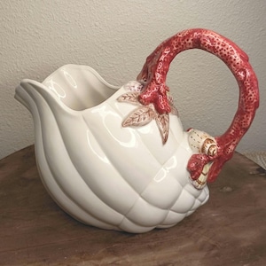 May include: White ceramic pitcher with a ribbed design and a red handle. The handle has a textured, branch-like appearance. Decorative leaves and shell accents adorn the pitcher's body. A unique and elegant piece for serving.