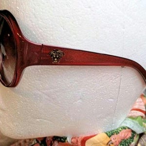 May include: Red-framed sunglasses with dark lenses. The sunglasses feature a gold-colored emblem on the temple. The frame has a slight sparkle. The sunglasses are a fashion accessory.