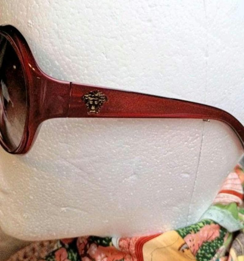 May include: Red-framed sunglasses with dark lenses. The sunglasses feature a gold-colored emblem on the temple. The frame has a slight sparkle. The sunglasses are a fashion accessory.