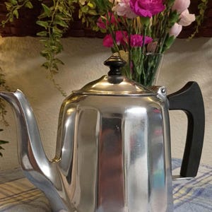 May include: A shiny, silver-colored metal teapot with a black handle and lid knob. The teapot is rectangular with a curved spout. A bouquet of pink and white flowers sits in a glass vase behind the teapot.
